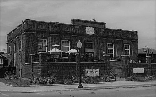 A historic brick building with a sign reading "Library," featuring outdoor seating and a lamppost in front.