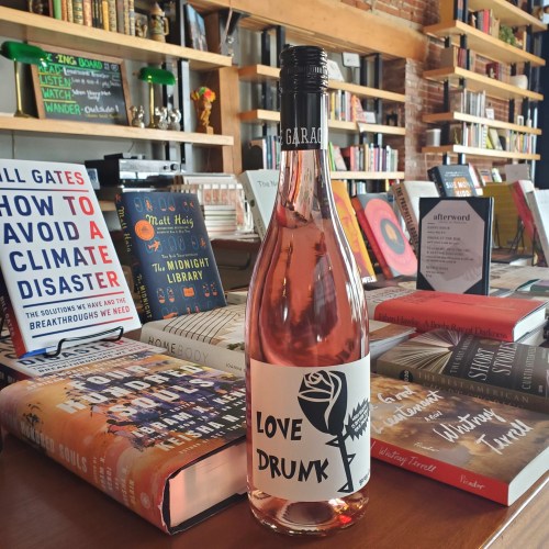 A bottle of rosé wine labeled "Love Drunk" in front of a display of various books on a wooden table.