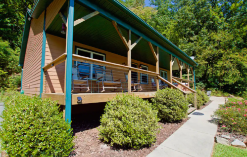 A cozy cabin with a wooden porch, surrounded by greenery and a pathway leading to the entrance.