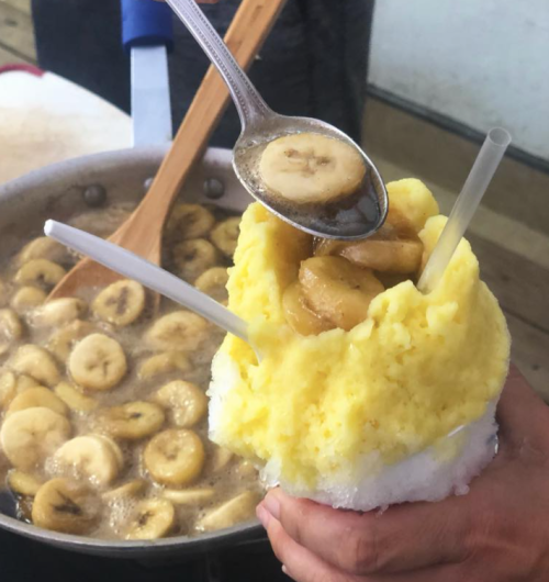 A hand holds a colorful snow cone topped with banana slices, with a pot of sliced bananas in syrup in the background.