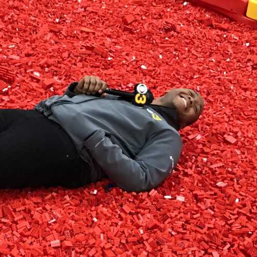 A person lies on a bed of red LEGO bricks, smiling and holding a microphone, in a playful setting.