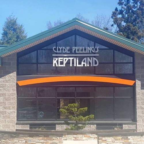 Sign for Clyde Peeling's Reptiland, a reptile zoo, with a modern building and trees in the background.