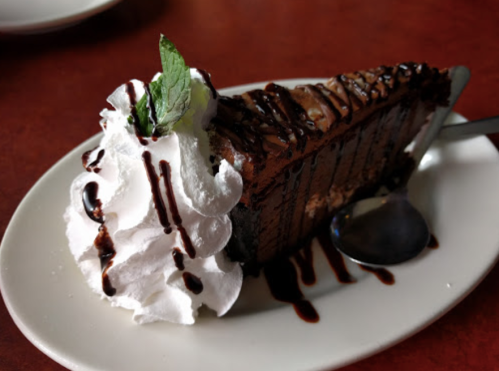 A slice of chocolate cake topped with whipped cream, chocolate drizzle, and a mint leaf on a white plate.