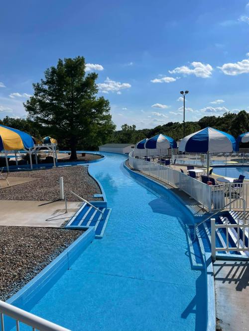 A winding blue water slide surrounded by umbrellas and trees under a clear blue sky.