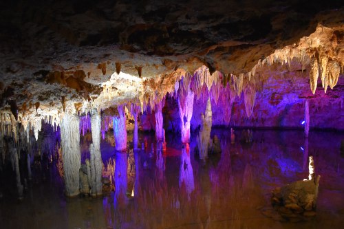 A colorful cave interior with stalactites and stalagmites reflected in still water, illuminated by vibrant lights.
