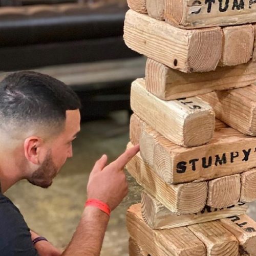 A person points at a wooden block tower labeled "STUMPY" in a casual indoor setting.