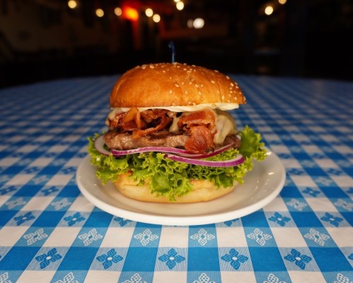 A delicious burger with bacon, cheese, lettuce, and onion on a sesame seed bun, served on a blue checkered tablecloth.