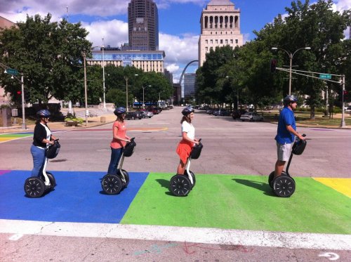 Four people riding Segways across a colorful crosswalk in an urban area, with trees and buildings in the background.