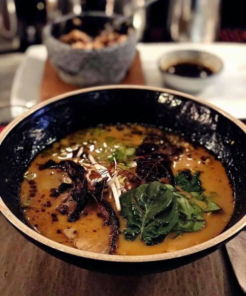 A bowl of rich ramen soup topped with greens, mushrooms, and herbs, with a side of dipping sauce in the background.