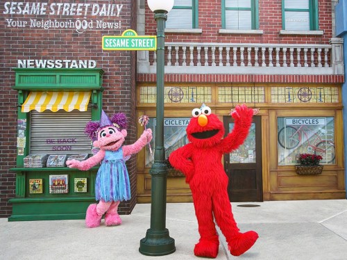 Elmo and Abby Cadabby pose playfully on Sesame Street near a newsstand and bicycle shop.