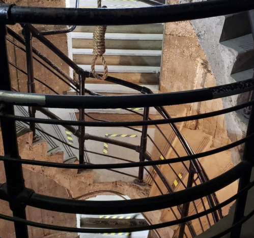 A spiral staircase with a rope hanging above, viewed through a metal railing. The walls are textured and weathered.