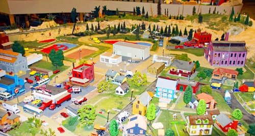 A detailed model train layout featuring buildings, vehicles, and trees in a miniature town setting.