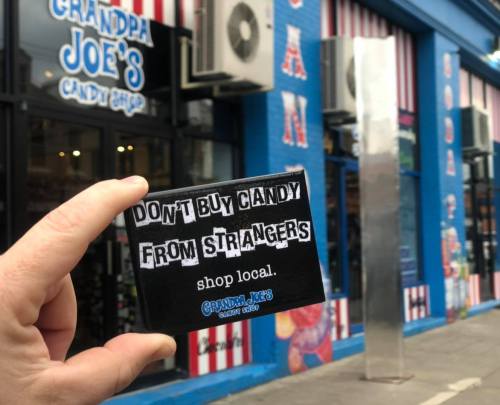 A hand holds a sign reading "Don't buy candy from strangers. Shop local." in front of Grandpa Joe's Candy Shop.
