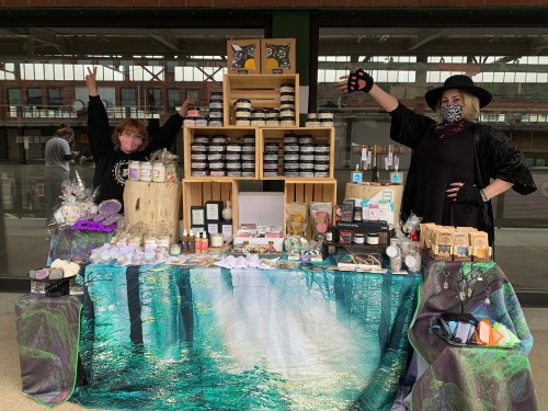 Two vendors at a market stand behind a colorful display of handmade products, smiling and posing with their arms raised.
