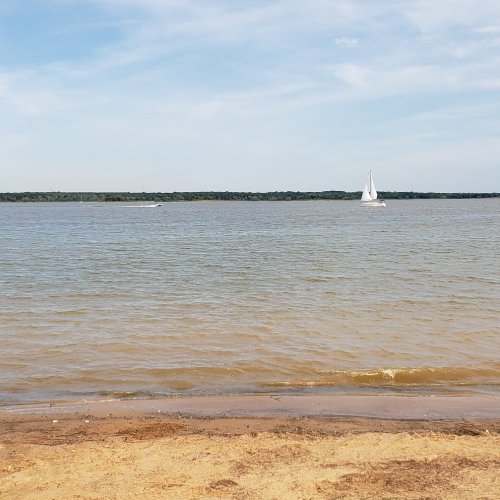 A serene view of a sandy beach with calm water and a sailboat in the distance under a clear blue sky.