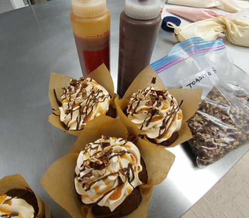 Four decorated cupcakes with frosting and drizzled sauces, alongside squeeze bottles and a bag of toppings.