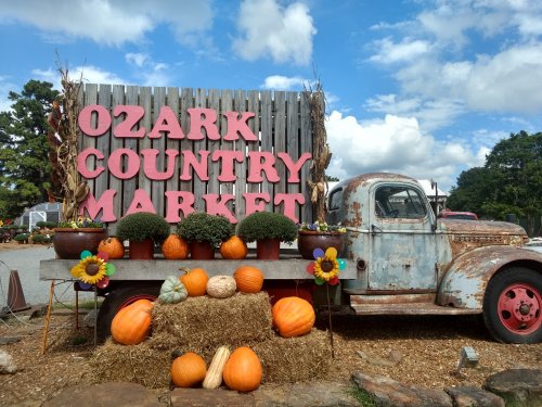 A vintage truck beside a sign reading "Ozark Country Market," decorated with pumpkins and sunflowers against a blue sky.