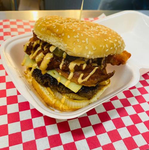 A stacked burger with bacon, lettuce, and sauce on a sesame seed bun, served in a takeout container with a checkered pattern.