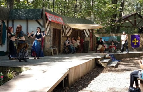 A lively outdoor performance with dancers and musicians on a wooden stage, surrounded by trees and rustic buildings.