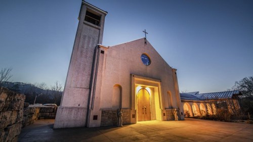A church building at dusk, illuminated from within, with a cross on top and a bell tower.