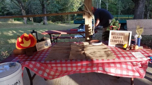 A picnic table with a red checkered tablecloth, decorations, and a sign welcoming campers in a wooded area.