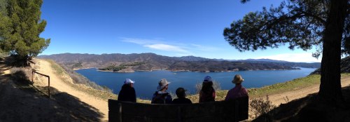 A group of five people sits on a bench overlooking a serene lake and mountains under a clear blue sky.