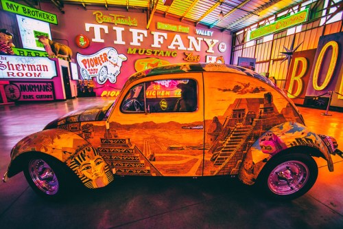 A colorful, artistic car with Egyptian motifs parked in a vibrant, neon-lit indoor space filled with signs.