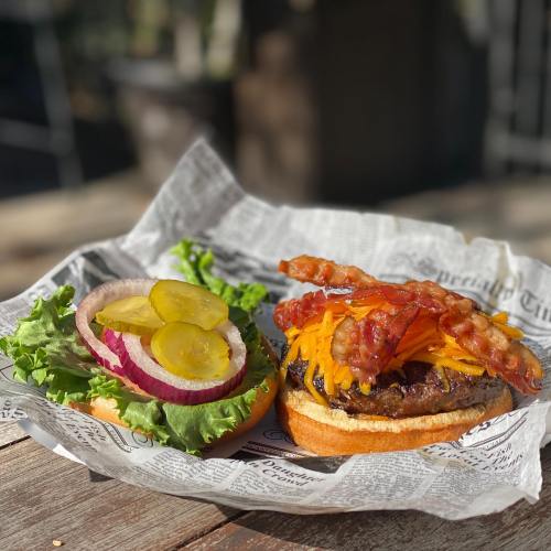 A delicious burger topped with bacon and cheese, served with lettuce, onion, and pickles on a newspaper-lined plate.