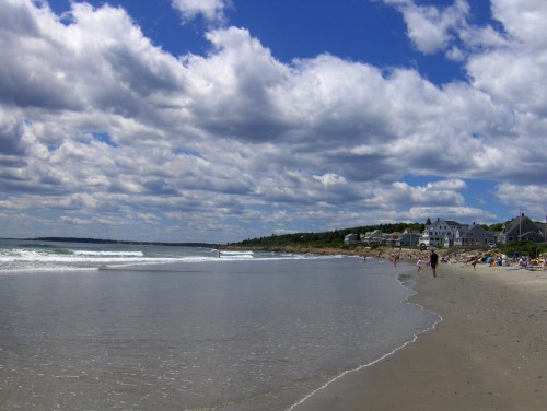 A sandy beach with gentle waves, under a cloudy sky, and a distant shoreline with beachgoers and houses.