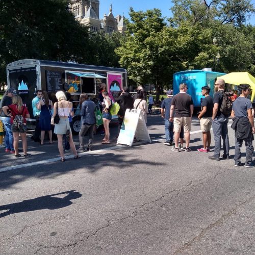A crowd gathers around food trucks at an outdoor event, with people waiting in line and enjoying the sunny day.