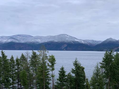 A serene lake surrounded by evergreen trees and snow-capped mountains under a cloudy sky.
