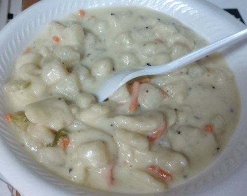 A bowl of creamy chicken and dumpling soup with carrots and herbs, served with a white plastic spoon.