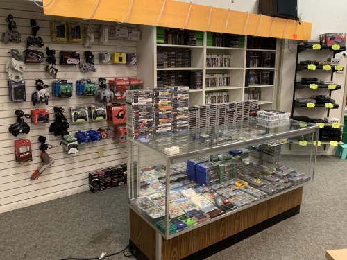 A retro video game store featuring shelves of games, consoles, and accessories, with a display case in the center.