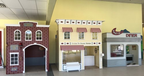 Three colorful playhouses: a brick building, a grocery market, and a diner, set in a bright indoor space.