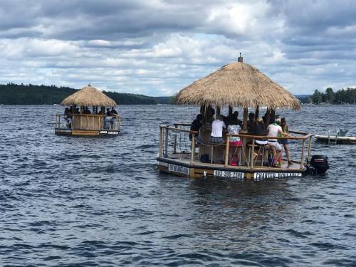 Two floating tiki huts on a lake, with people enjoying the water under a cloudy sky.