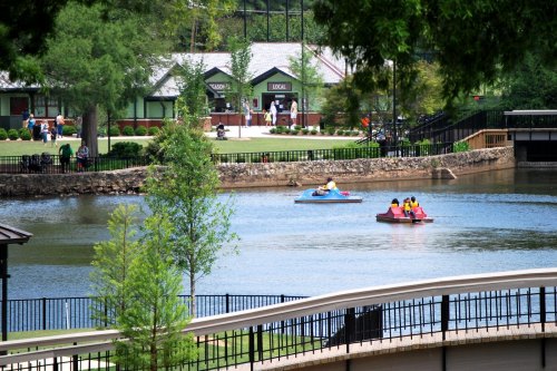 Two paddle boats on a calm lake, with a park and buildings in the background on a sunny day.
