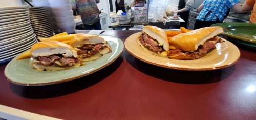 Two plates with large sandwiches filled with meat and toppings, accompanied by orange fries in a casual dining setting.