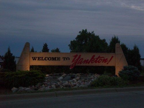 Sign welcoming visitors to Yankton, surrounded by trees and rocks, under a cloudy evening sky.