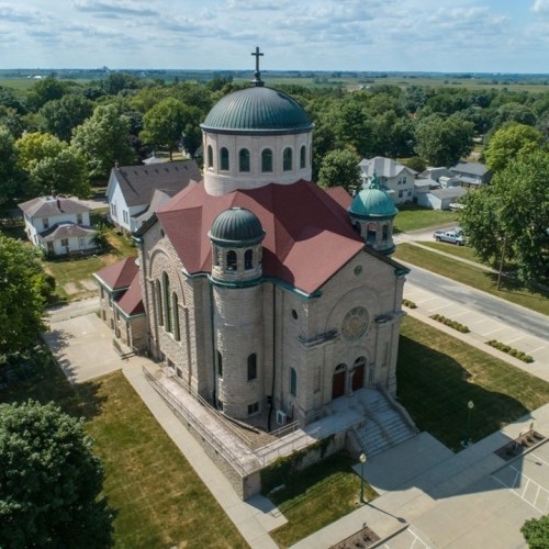 Aerial view of a large stone church with a red dome and cross, surrounded by green trees and residential buildings.