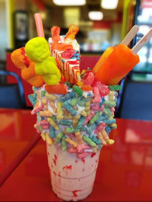 A colorful milkshake topped with candy, gummies, and sweets, served in a cup decorated with vibrant cereal pieces.