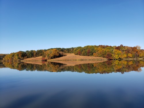 A serene lake reflecting a colorful autumn landscape with trees in shades of orange, yellow, and green under a clear blue sky.