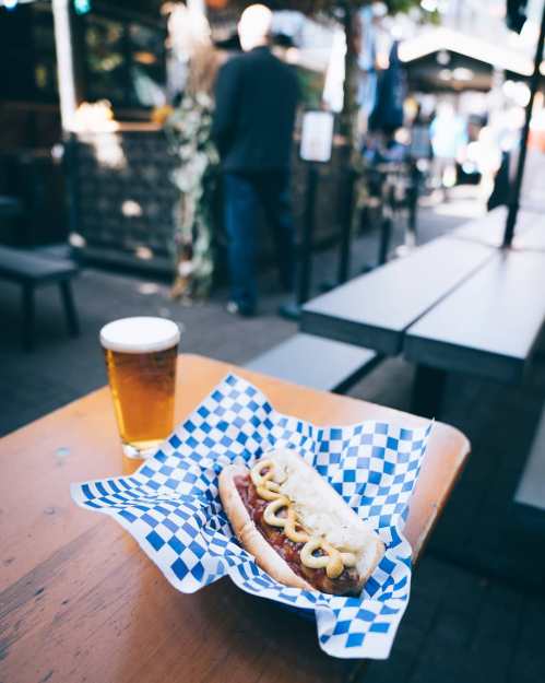 A hot dog with mustard on a blue checkered plate, next to a glass of beer, set on a wooden table outdoors.