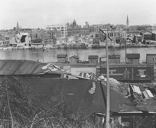 Black and white photo of a riverfront cityscape with buildings, warehouses, and a train near the water's edge.