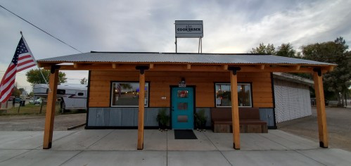 A small wooden building with a metal roof, featuring a blue door and an American flag outside.