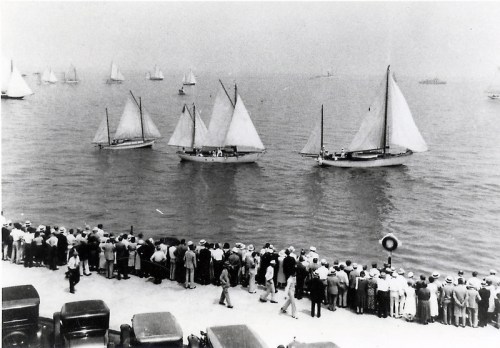 A black and white photo of a crowd watching sailboats on a calm waterway, with vintage cars parked nearby.