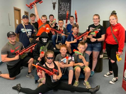 A group of kids and teens posing with toy guns in a playful setting, some wearing sunglasses and costumes.
