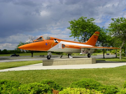 An orange jet aircraft on display, mounted on a pedestal, surrounded by grass and trees under a cloudy sky.