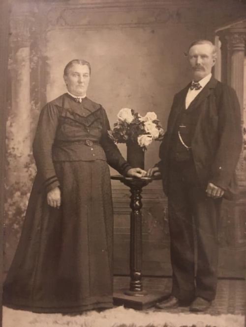 A vintage black-and-white photo of a man and woman standing beside a floral arrangement on a pedestal.
