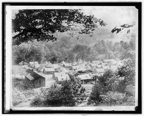 Aerial view of a campsite with multiple tents surrounded by trees and a wooden building in the foreground.