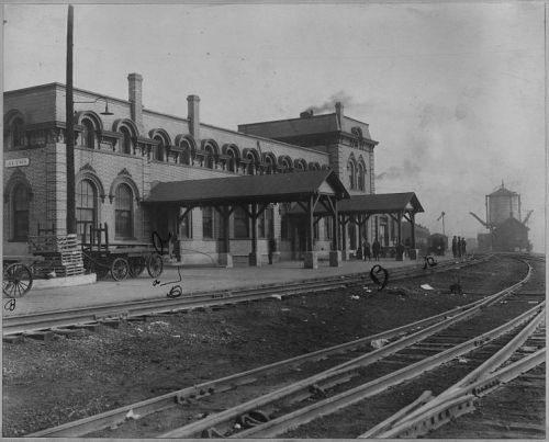 Historic train station with brick architecture, horse-drawn carts, and railway tracks leading into the distance.